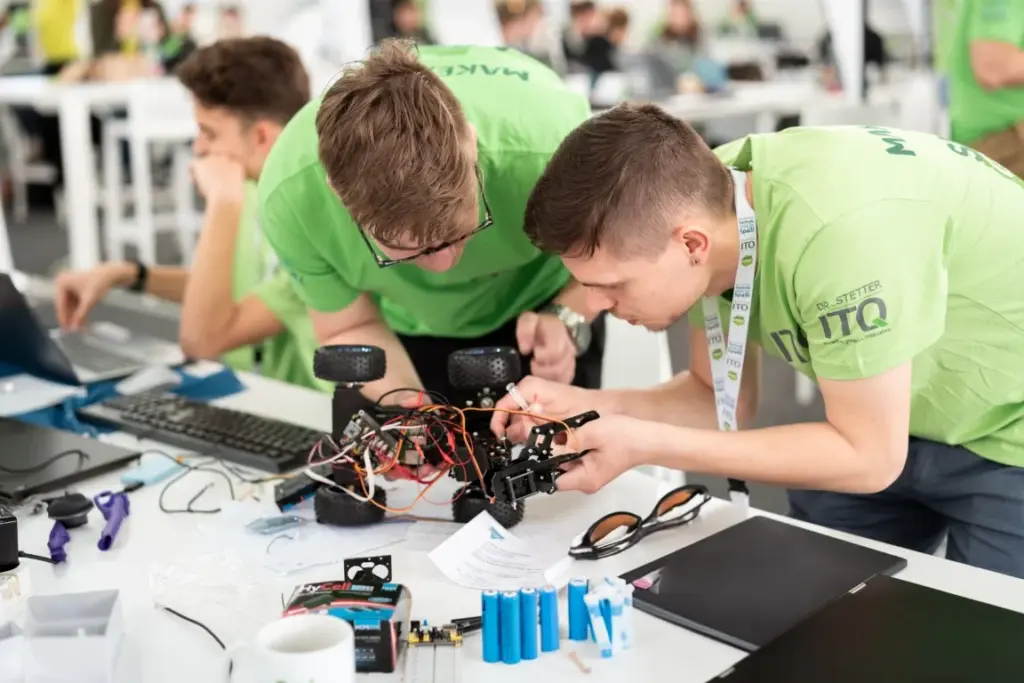 Zwei junge Männer in grünen Hemden bauen an einem Arbeitstisch gemeinsam einen kleinen Roboter auf Rädern zusammen und begutachten ihn, umgeben von Werkzeugen, Elektronikbauteilen, Laptops und Papieren.