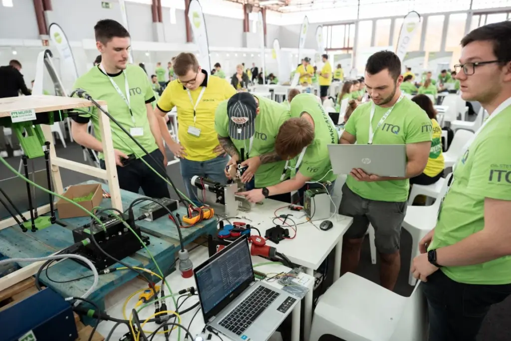 Eine Gruppe von Menschen in grünen Hemden arbeitet während einer Technologieveranstaltung an einem Tisch mit Laptops, Kabeln und Geräten an Elektronik.