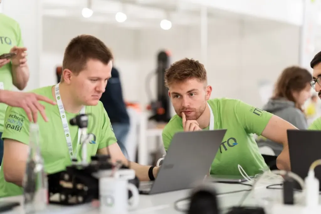 Zwei Männer in grünen Hemden konzentrieren sich auf einen Laptop an einem Tisch, auf dem elektronische Geräte, Tassen und Schlüsselbänder stehen, in einem hellen, modernen Arbeitsbereich.
