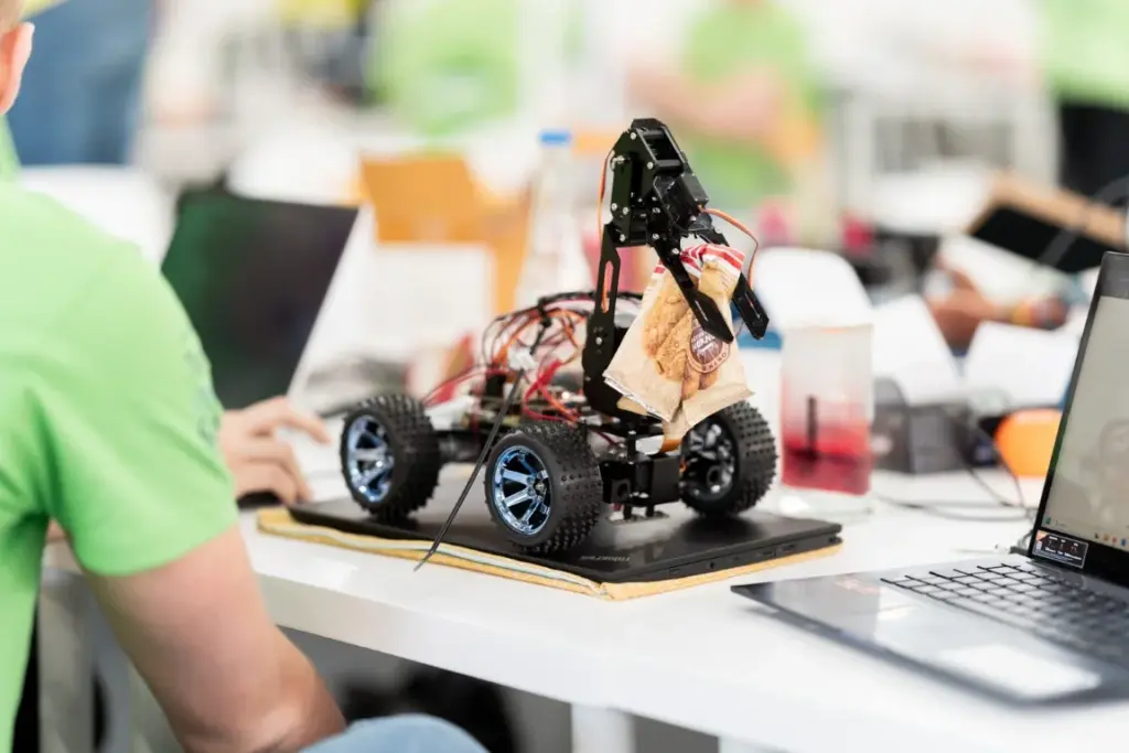 Ein kleines Roboterfahrzeug mit blauen Rädern und freiliegenden Kabeln steht auf einem Schreibtisch neben einem Laptop; ein Greifarm hält ein Stück Pappe fest.