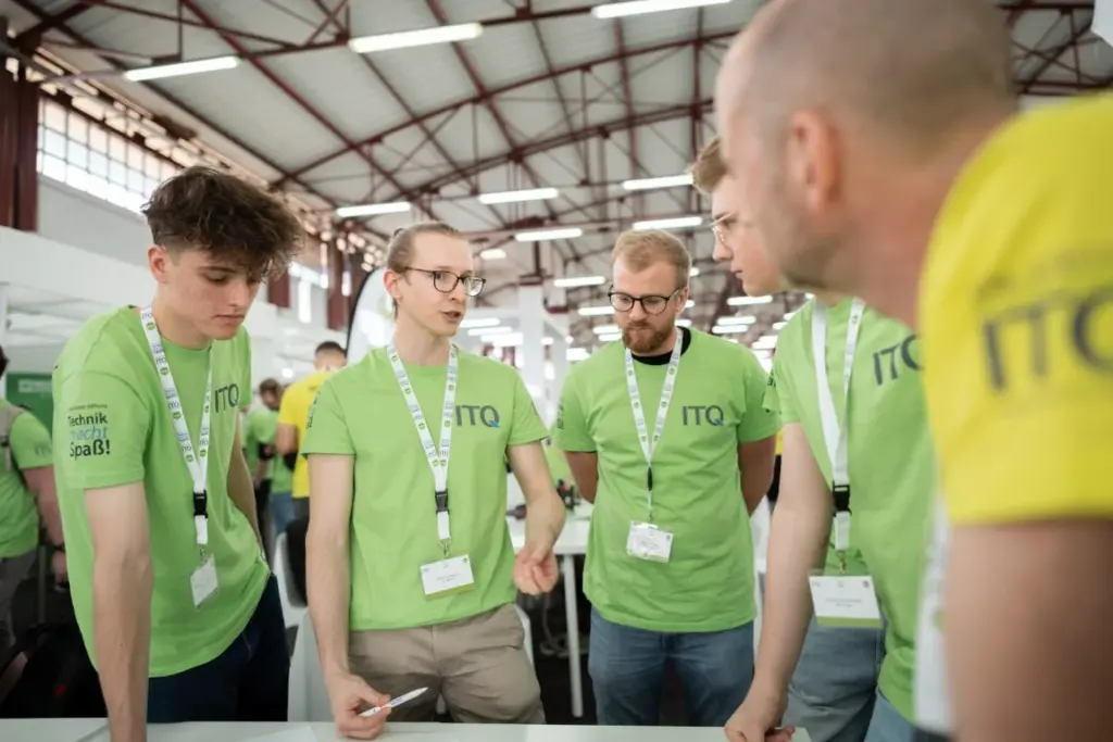 Fünf Personen, die ITQ-Shirts und Namensschilder tragen, stehen in einer Gruppe in einem Innenraum und führen unter einer hohen Decke mit sichtbaren Balken ein intensives Gespräch.