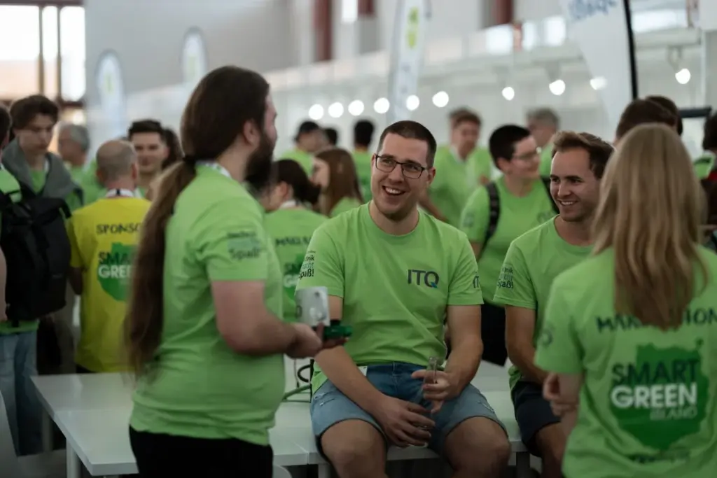 Eine Gruppe von Menschen in einheitlichen grünen T-Shirts unterhält sich und lächelt bei einer Veranstaltung in einem Saal, im Hintergrund sind Tische und Banner zu sehen.