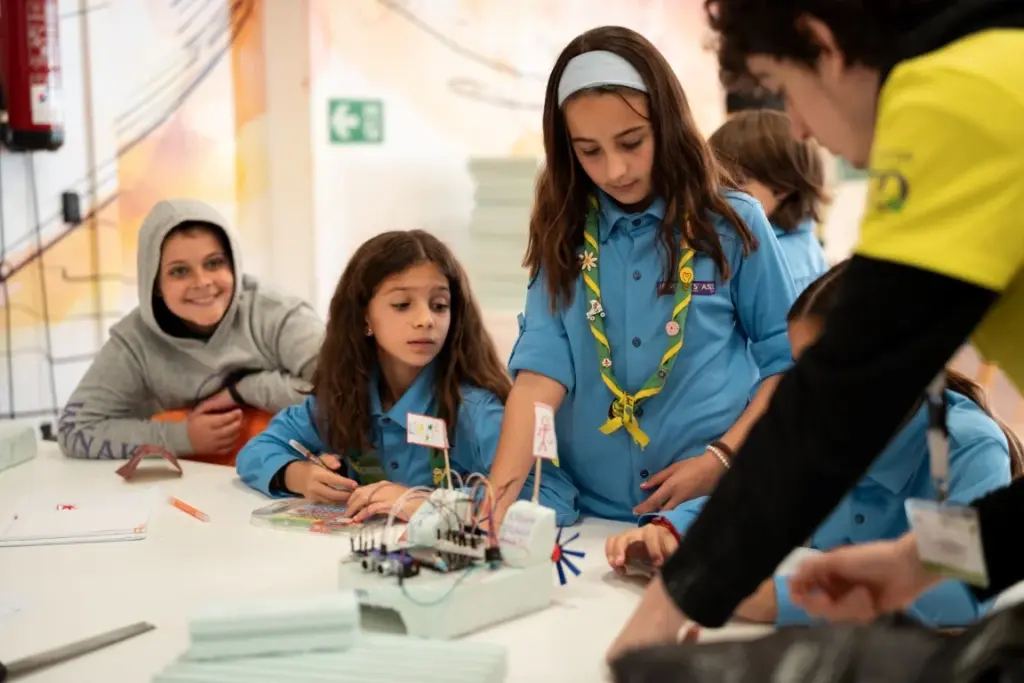 Kinder in blauen Uniformen arbeiten an einem Tisch an einem Elektronikprojekt, unterstützt von einem Erwachsenen in einem gelben Hemd, in einem Klassenzimmer.