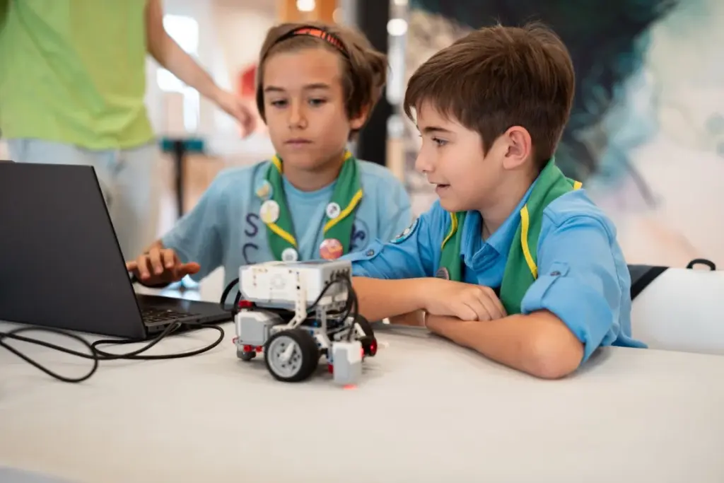 Zwei Kinder in Schuluniformen sitzen an einem Tisch und programmieren mithilfe eines Laptops einen kleinen Roboter auf Rädern. Beide sind ganz auf den Bildschirm konzentriert.