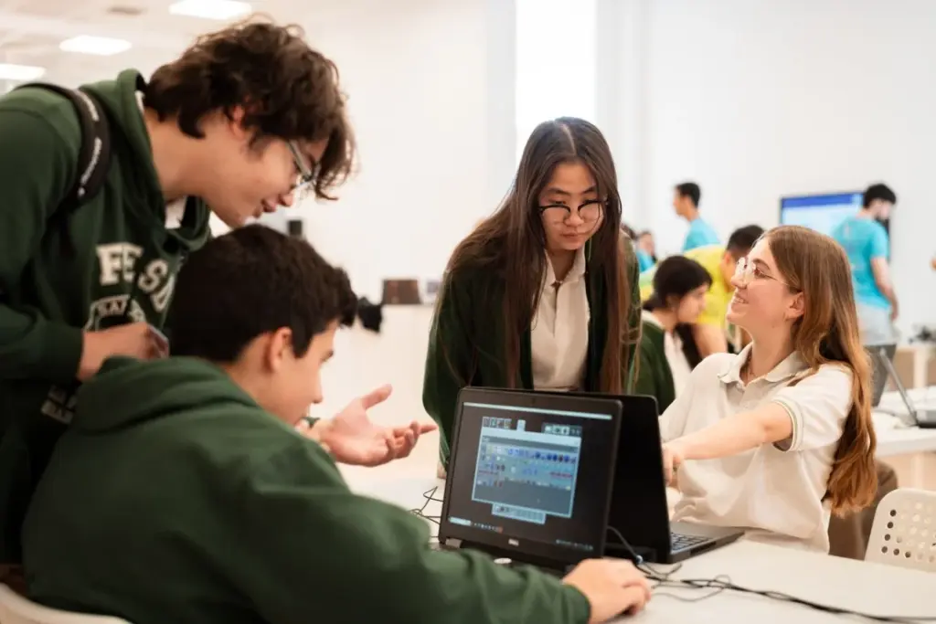 Vier Schüler versammeln sich in einem Klassenzimmer um einen Laptop und diskutieren über etwas, das auf dem Bildschirm zu sehen ist, während andere im Hintergrund arbeiten.