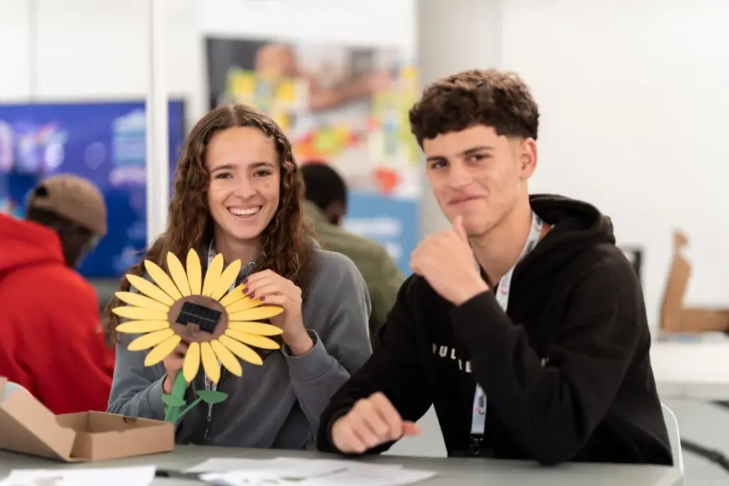 Zwei junge Menschen sitzen drinnen an einem Tisch. Einer hält einen sonnenblumenförmigen Gegenstand in der Hand, in dessen Mitte sich ein kleines Solarpanel befindet. Beide lächeln. Auf dem Tisch liegen Papiere und eine Schachtel.