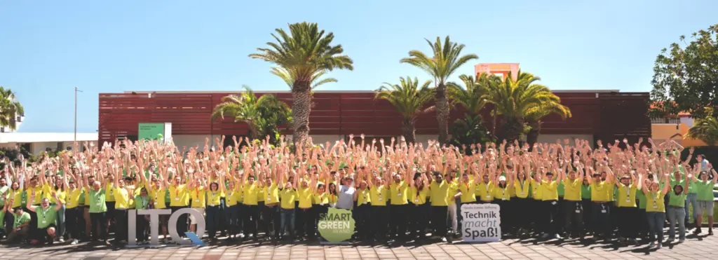 Eine große Gruppe von Menschen in gelben T-Shirts steht draußen unter Palmen und hebt gleichzeitig die Arme. Einige halten Schilder mit den Aufschriften „TQ“, „SMART GREEN“ und „Technik macht Spaß!“.