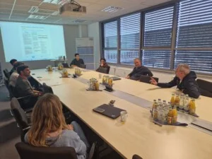 A group of people are sitting around a conference table with drinks, whilst one person is speaking and a document is being projected onto a screen.