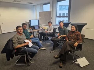 Five people are sitting in a modern office, gathered in a semi-circle with laptops and documents, and appear to be holding a meeting or discussion. A screen and a whiteboard can be seen in the background.