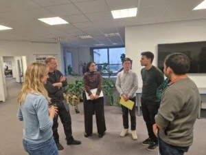 Six people are standing in a circle in an office, chatting and holding papers in their hands; desks and plants can be seen in the background.