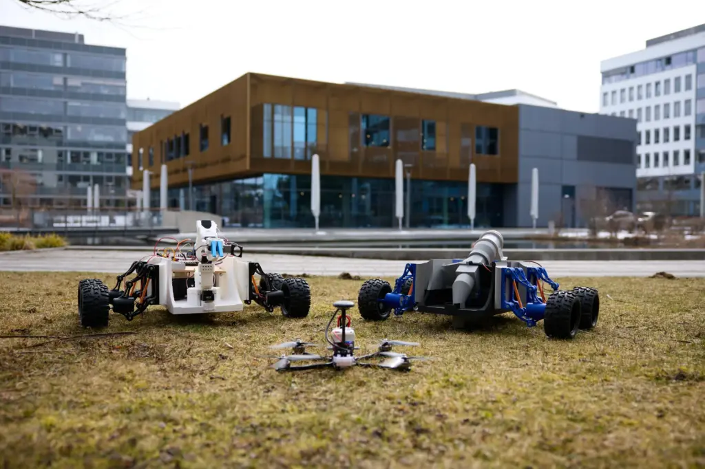 Zwei kleine Roboterfahrzeuge und eine Drohne stehen auf einer Rasenfläche vor einem modernen Gebäude mit großen Glasfenstern.