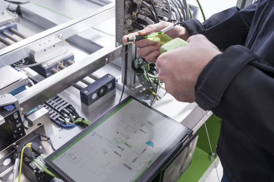 Eine Person schließt ein grünes Kabel an ein Industriegerät neben einem Touchscreen an, auf dem ein technisches Diagramm angezeigt wird; um sie herum befinden sich verschiedene Kabel und Maschinen.