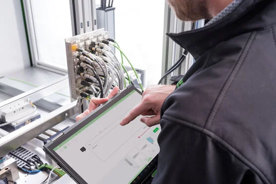 Eine Person, die eine dunkle Jacke trägt, bedient oder überwacht mithilfe eines Touchscreen-Tablets ein industrielles Bedienfeld, das über mehrere Kabel angeschlossen ist.